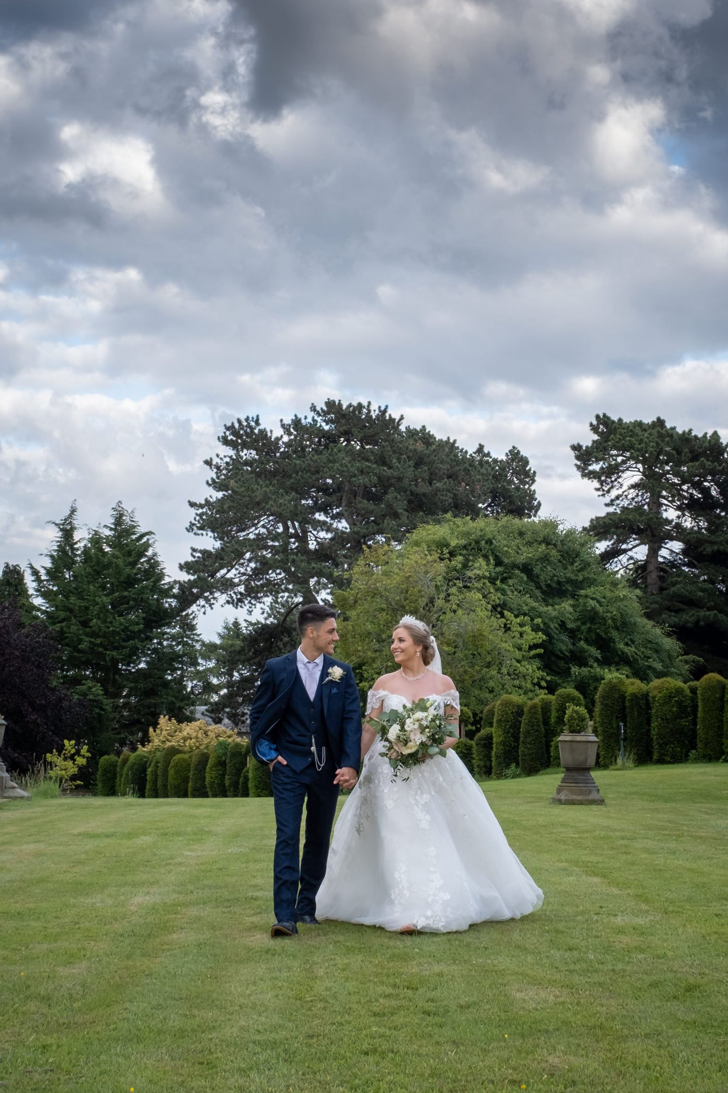 Wedding couple poses on lush green lawn with dramatic cloudy sky backdrop.