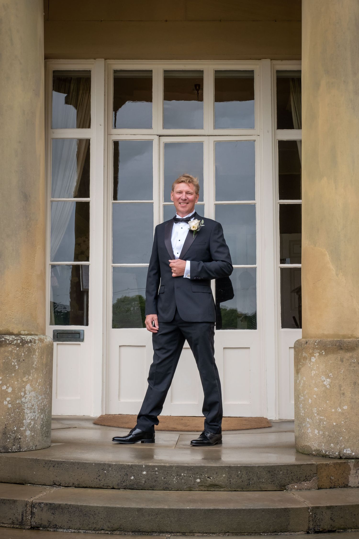 Two people in formal black tuxedos stand on stone steps in front of a grand entrance.