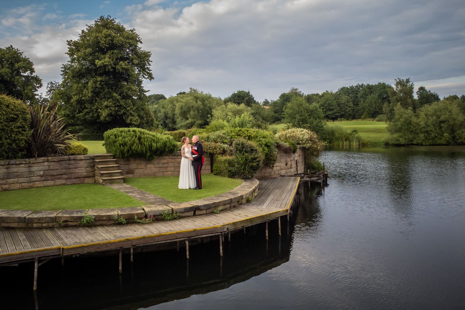 Wedding couple standing on curved stone pathway beside tranquil lake.