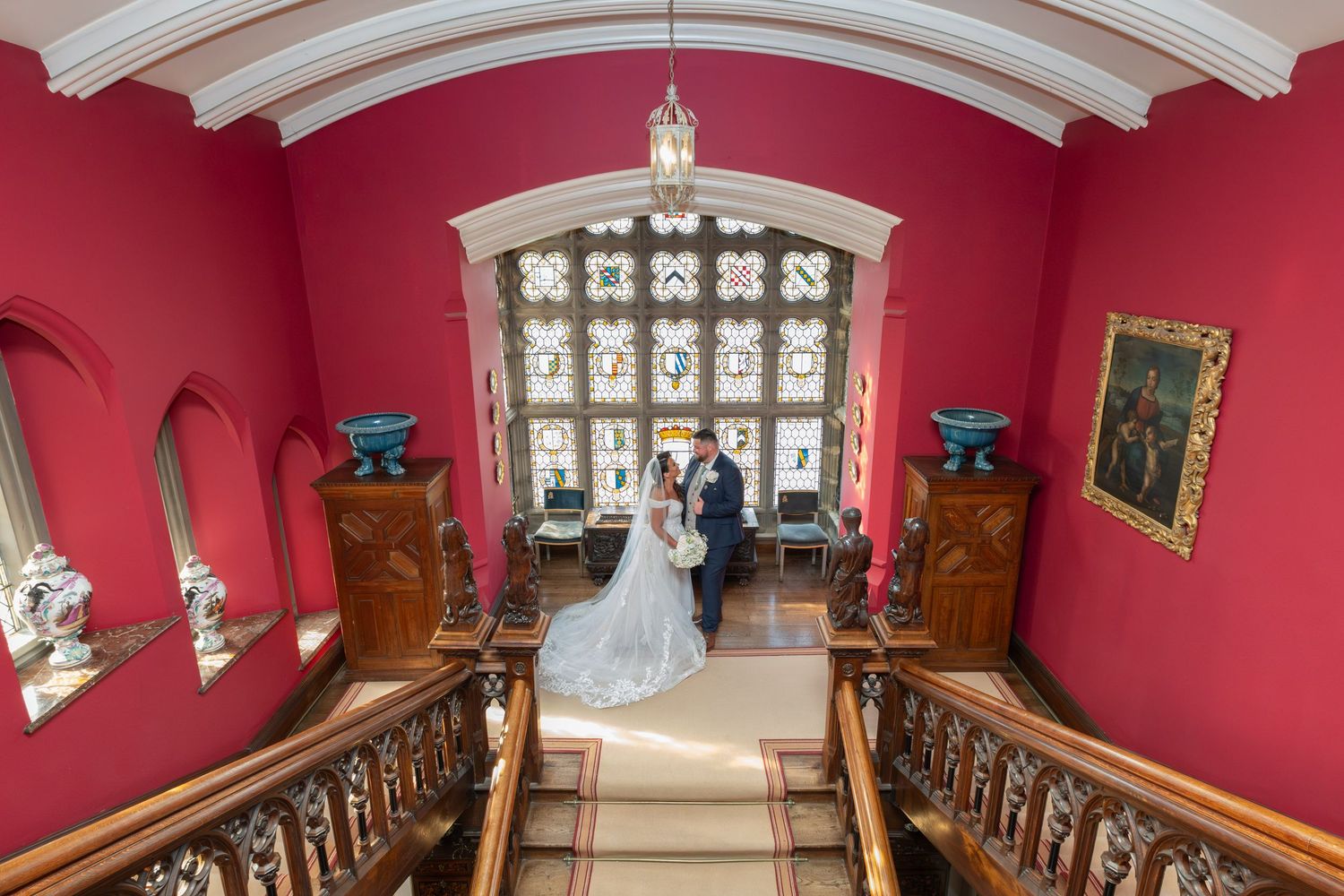 A wedding scene on an elegant red staircase with ornate woodwork and arched windows.