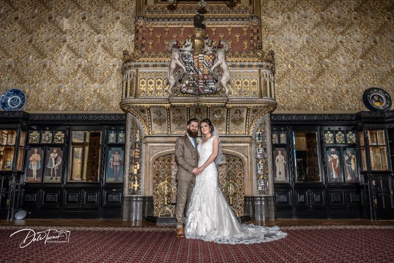 A couple poses in front of an ornate carved wooden wall in a historic venue.