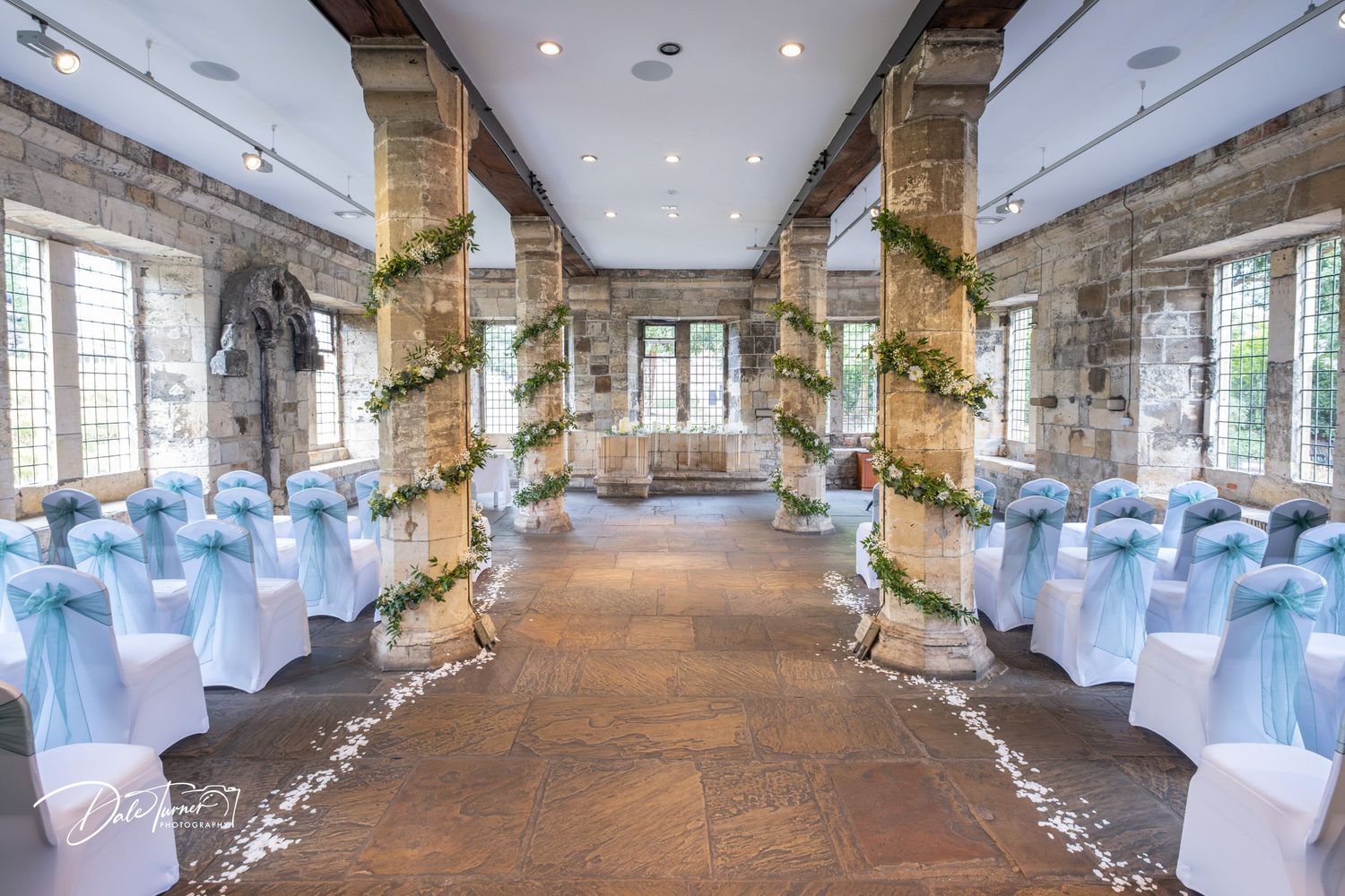 Wedding ceremony venue with blue chair covers and floral decorations along stone pillared aisle.