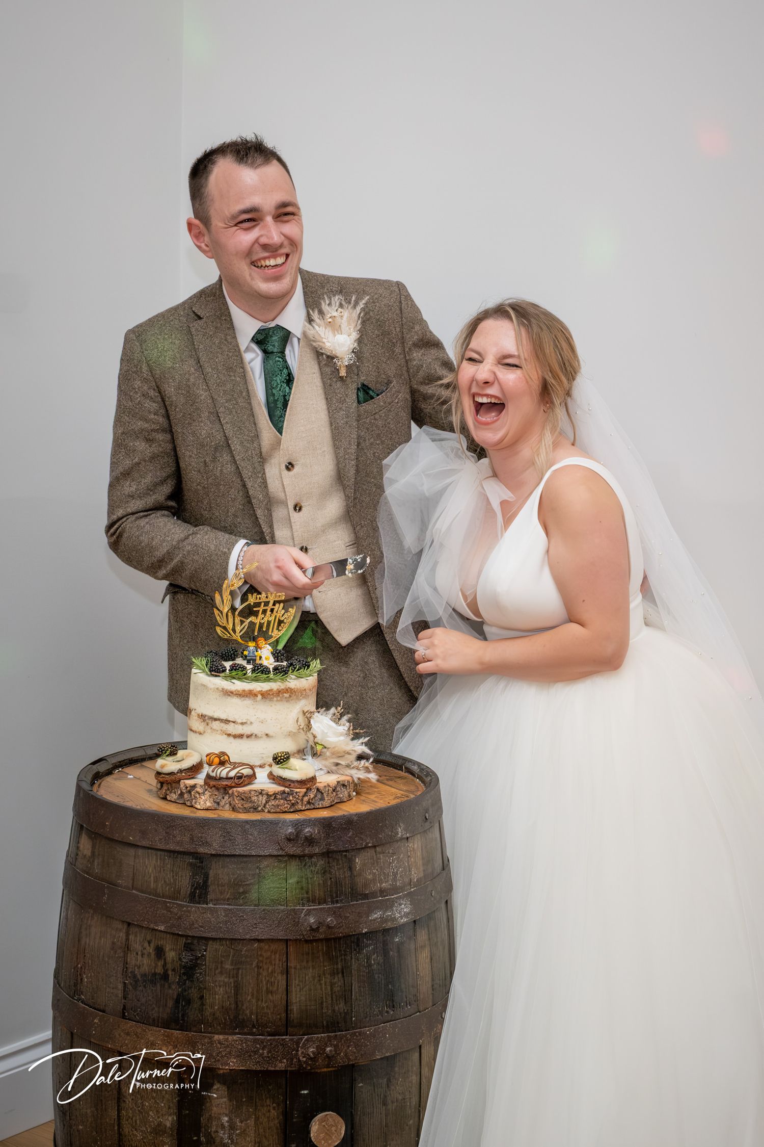 Wedding couple shares a joyful moment while cutting their cake displayed on a rustic wooden barrel.