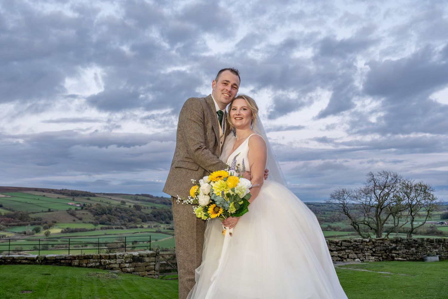 A couple embraces in front of a dramatic cloudy countryside landscape with rolling green hills in the background.
