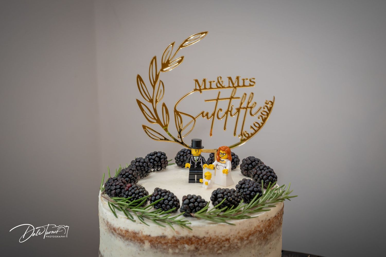 A white wedding cake decorated with fresh blackberries and delicate greenery sits elegantly against a grey background.