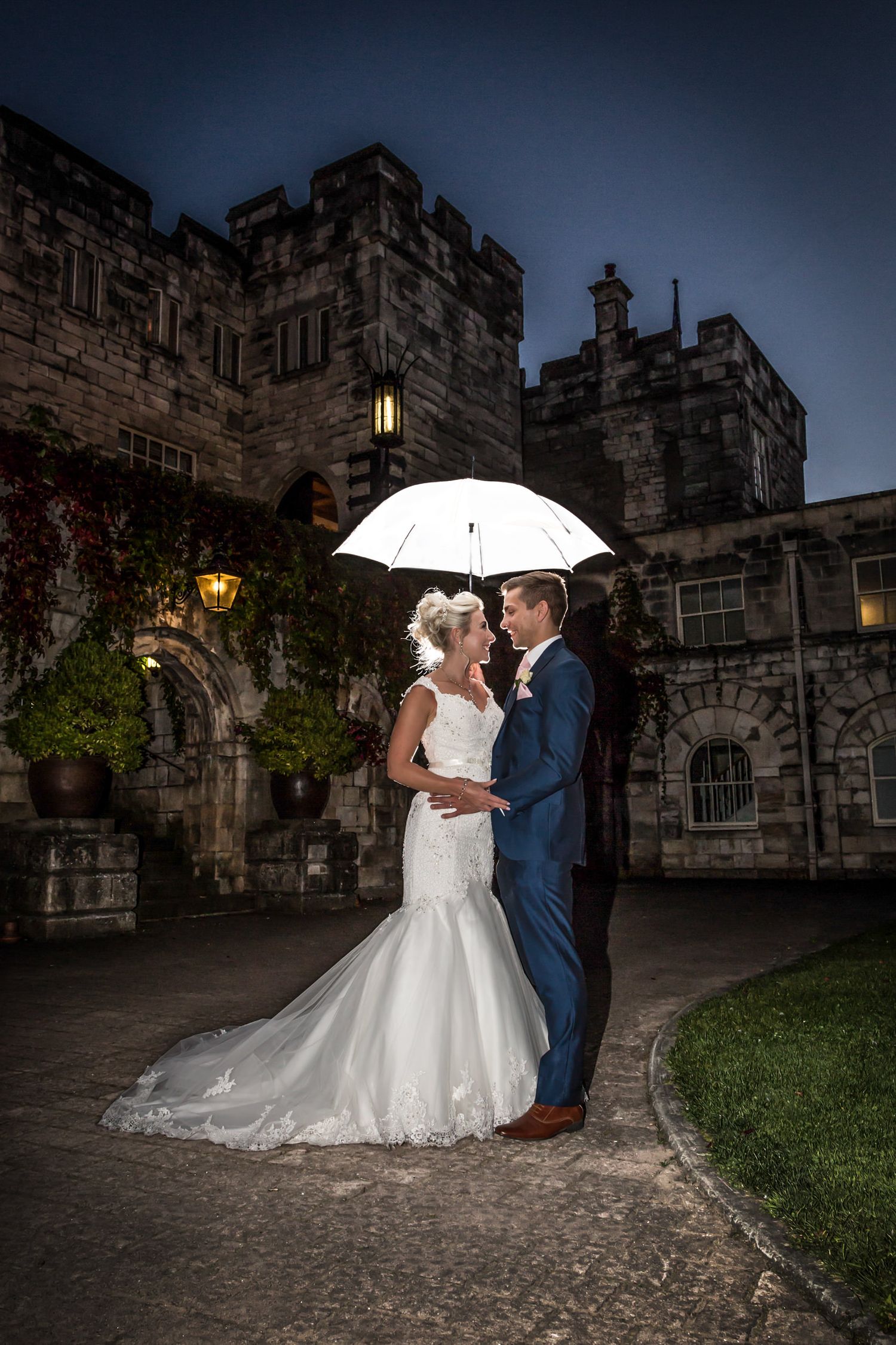 Evening wedding portrait with white umbrella against dramatic castle backdrop.