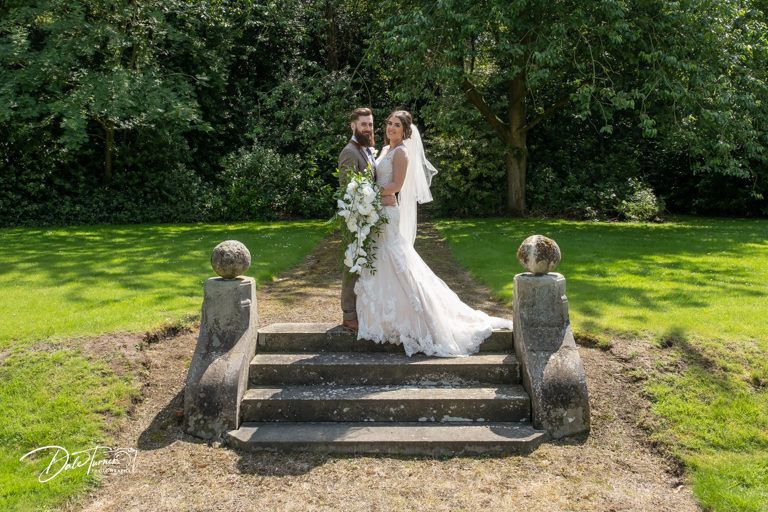 Wedding couple poses between decorative stone pillars in garden setting.