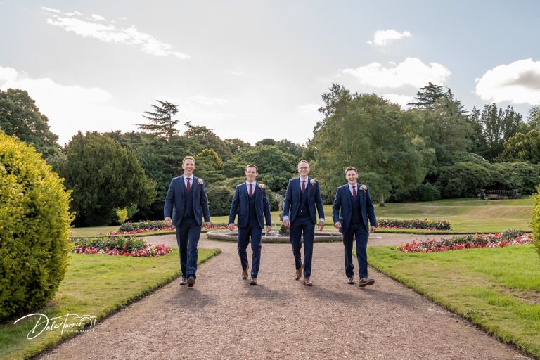 Four groomsmen in navy suits walking down a garden path surrounded by landscaped grounds.