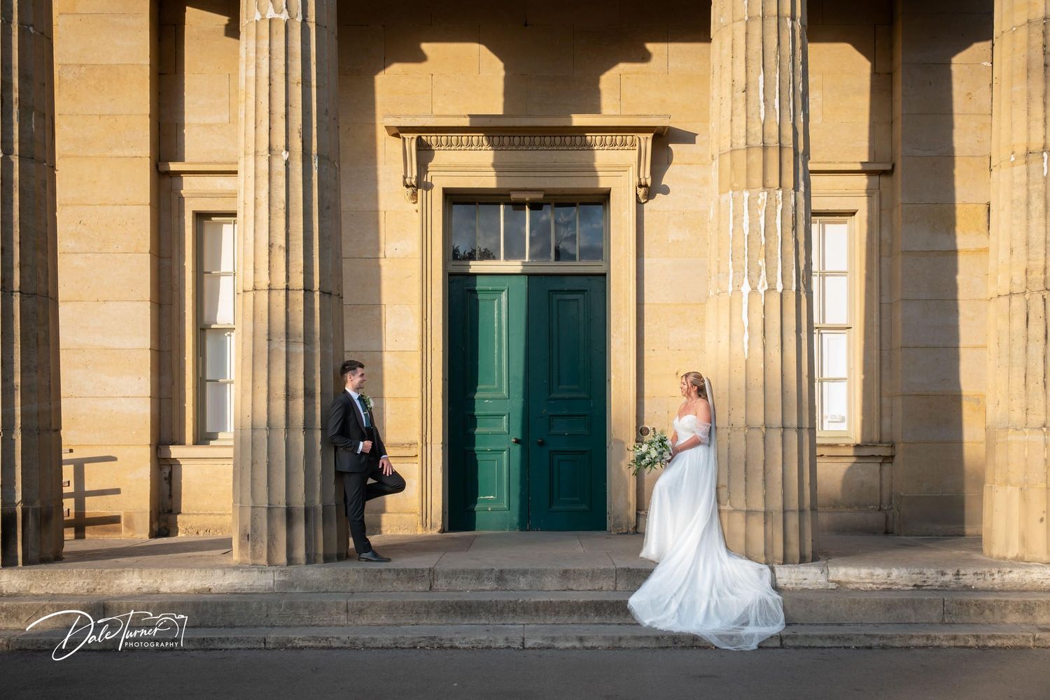 A couple stands between elegant stone columns in front of a green door at a historic venue.