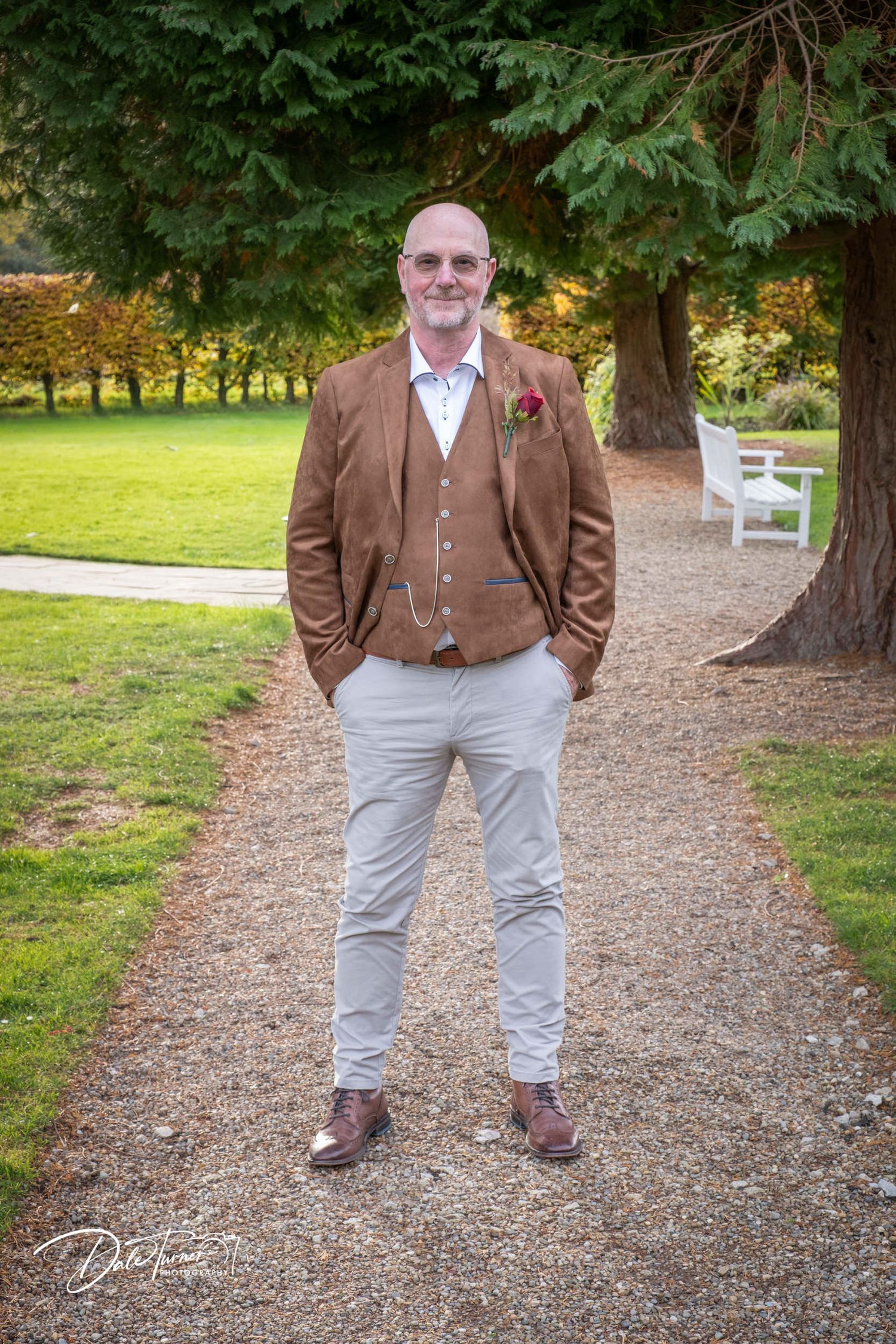 Figure in traditional brown tweed attire standing on gravel pathway under trees.