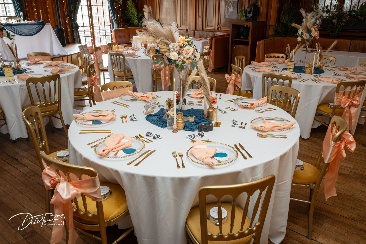 Elegant wedding reception table with gold chairs, white linens, pink plates, blue centerpiece, and floral arrangements in a decorated venue.