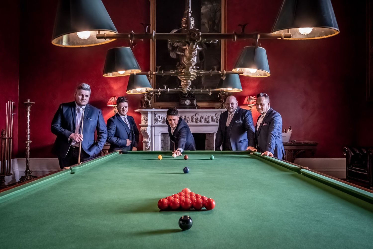 Snooker players gather around vintage pool table in classic red-walled billiards room.
