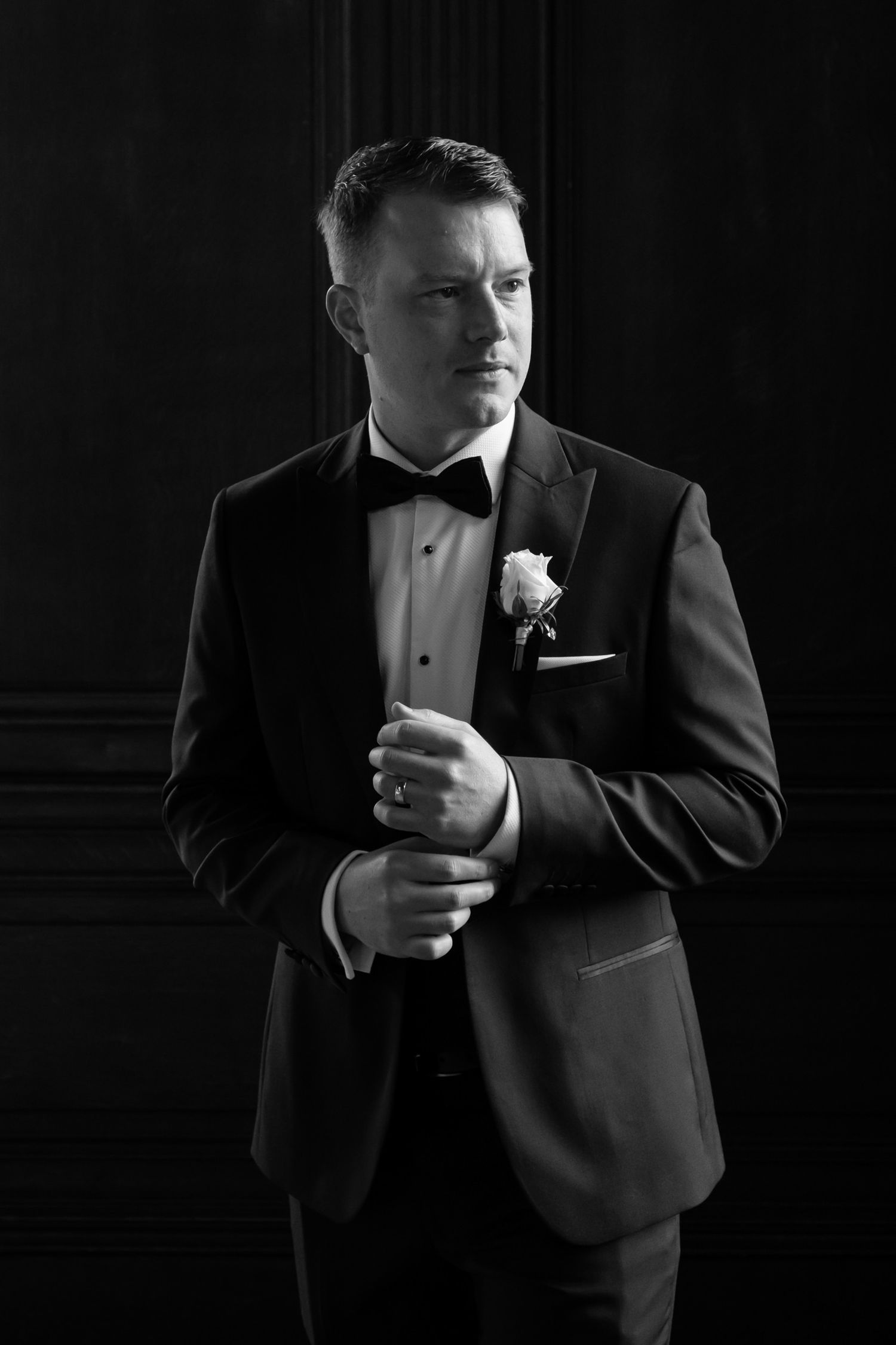 Close-up of a groom in black tuxedo adjusting his tie against dark background.