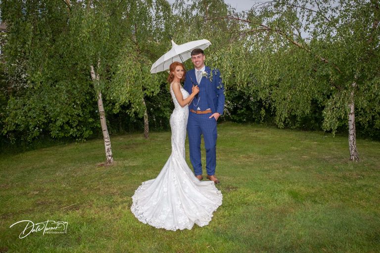 Romantic wedding couple embraces on grassy lawn surrounded by birch trees.