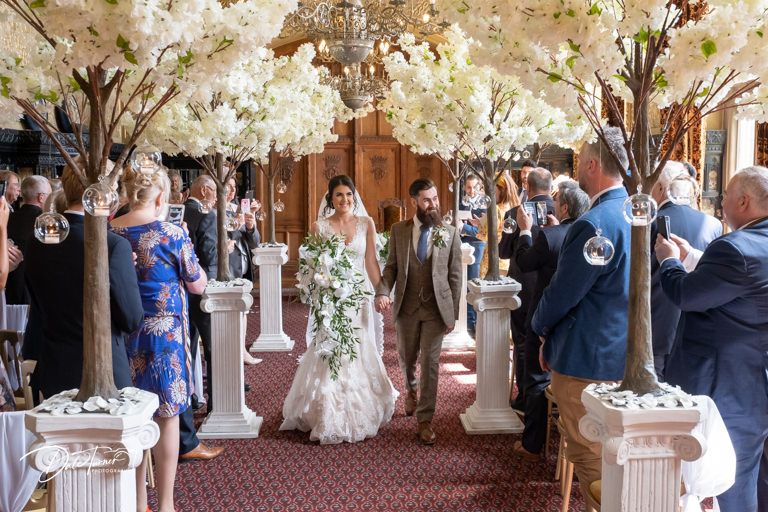 Wedding ceremony guests gathered in elegant hall with decorative white floral trees.