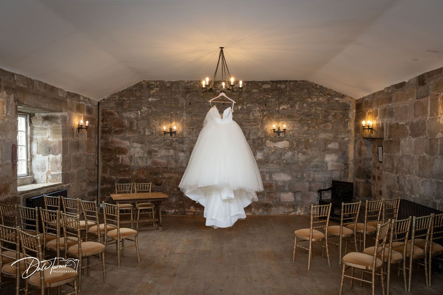 A white wedding dress hangs from a chandelier in a rustic stone room with wooden chairs arranged for a ceremony.