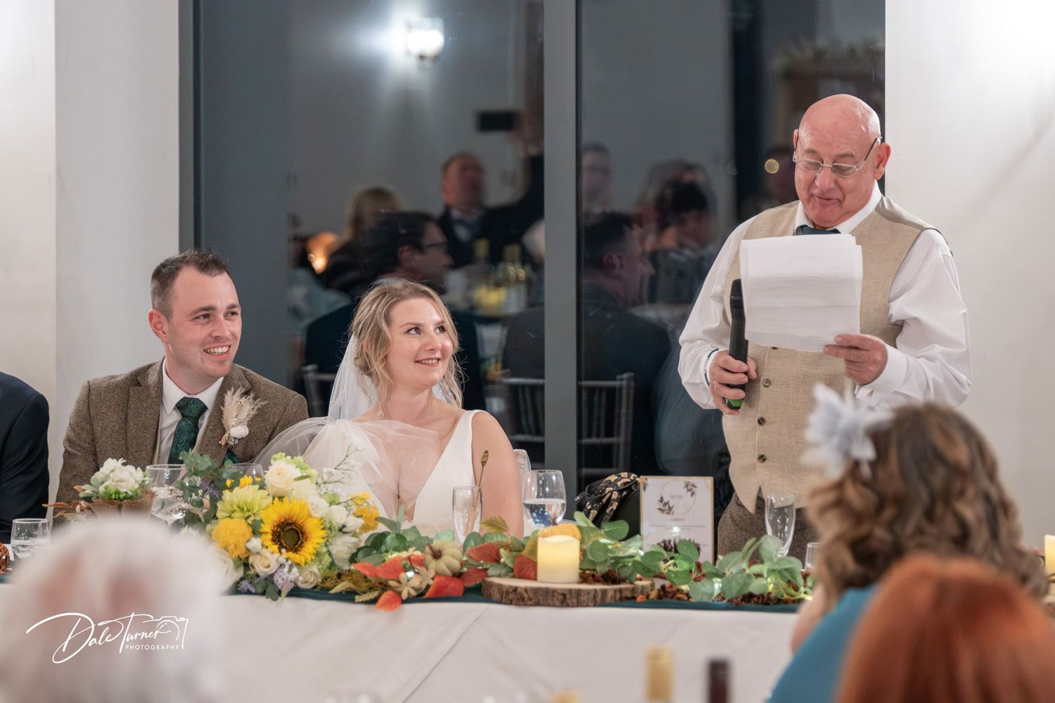 Wedding guests dining at elegant tables with yellow sunflower centerpieces in a bright reception venue.