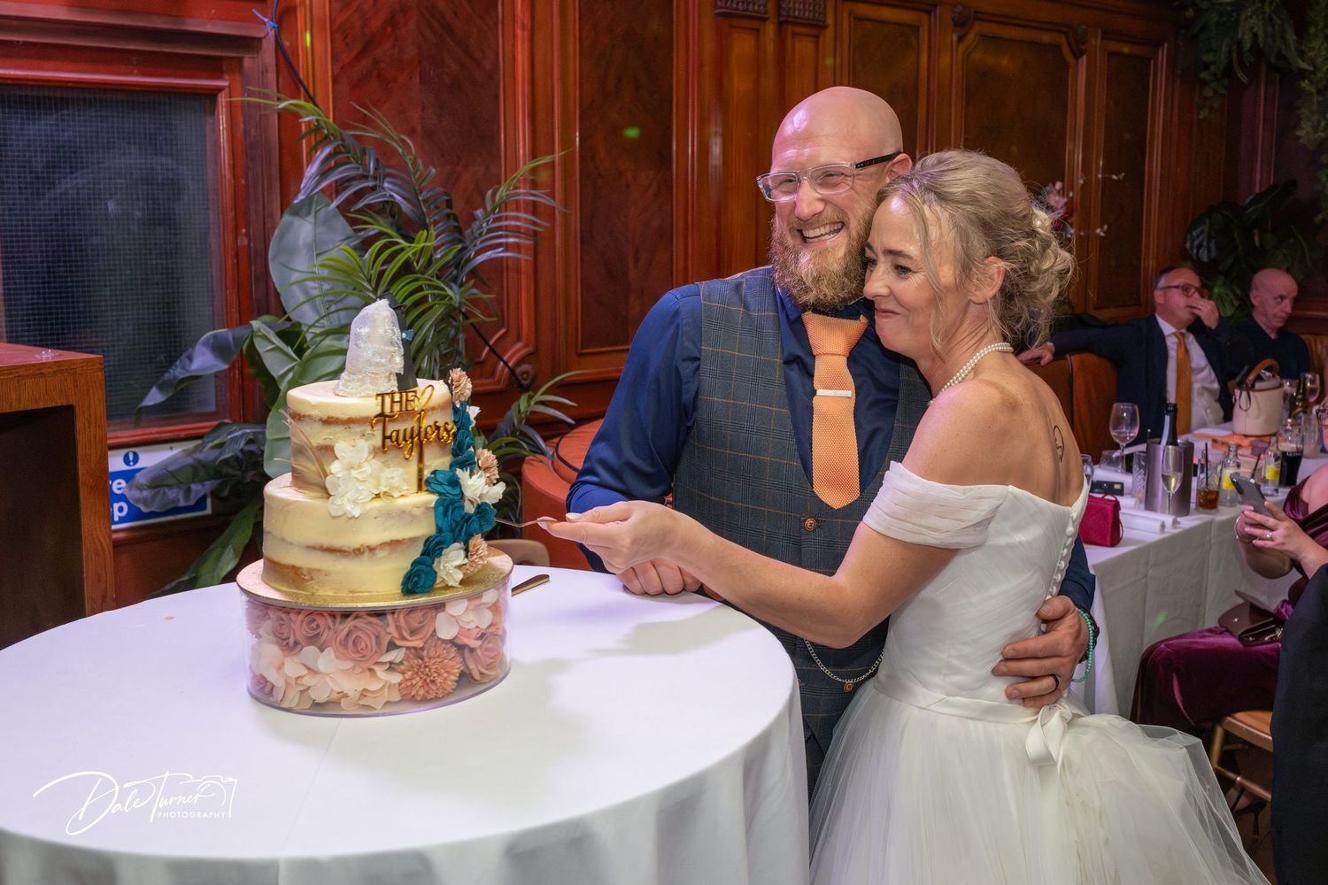 Bride and groom cutting their elegant tiered wedding cake at a wedding reception in a wood-paneled venue.