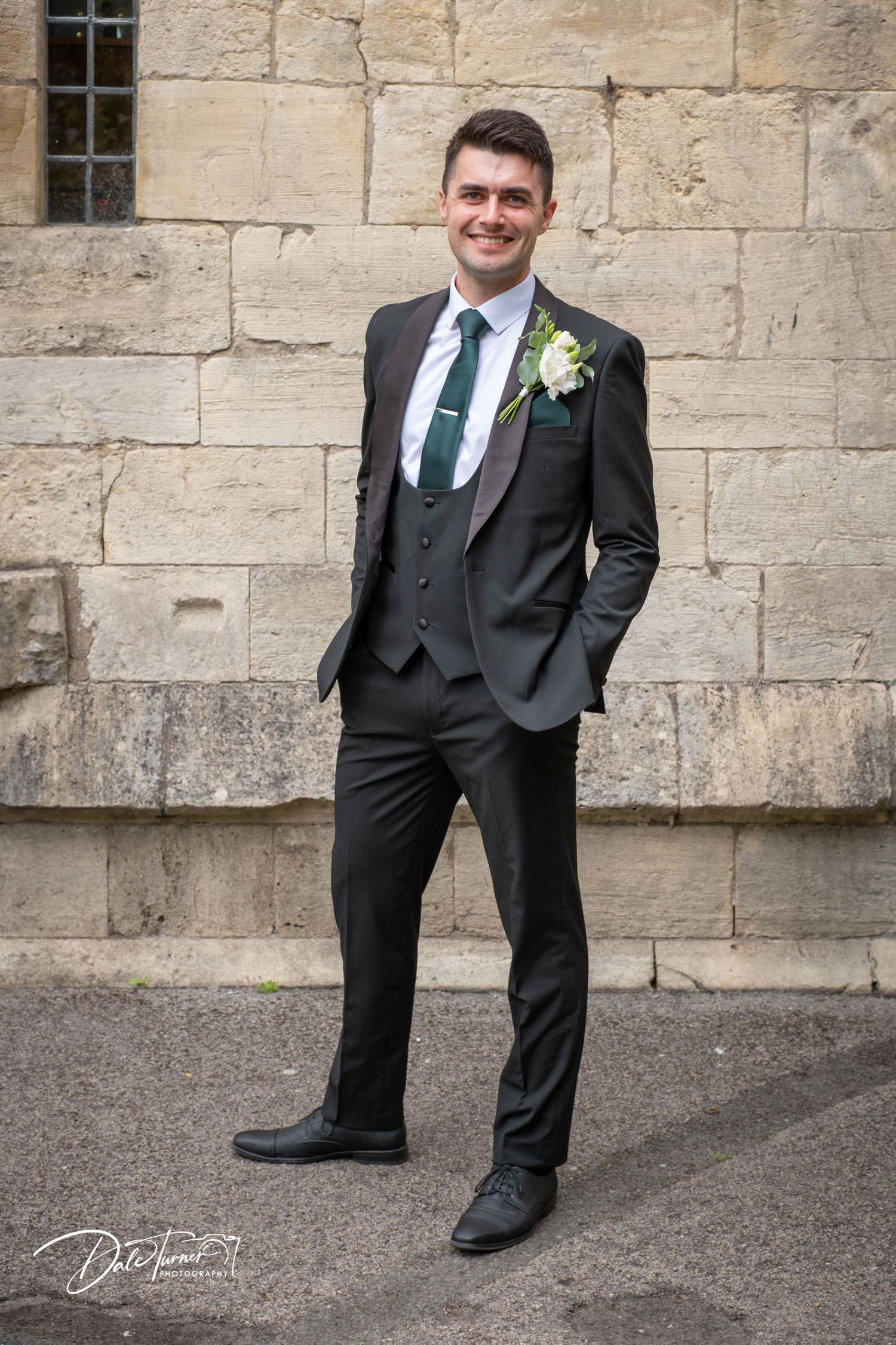 Full length portrait of groom in formal attire against stone building backdrop.