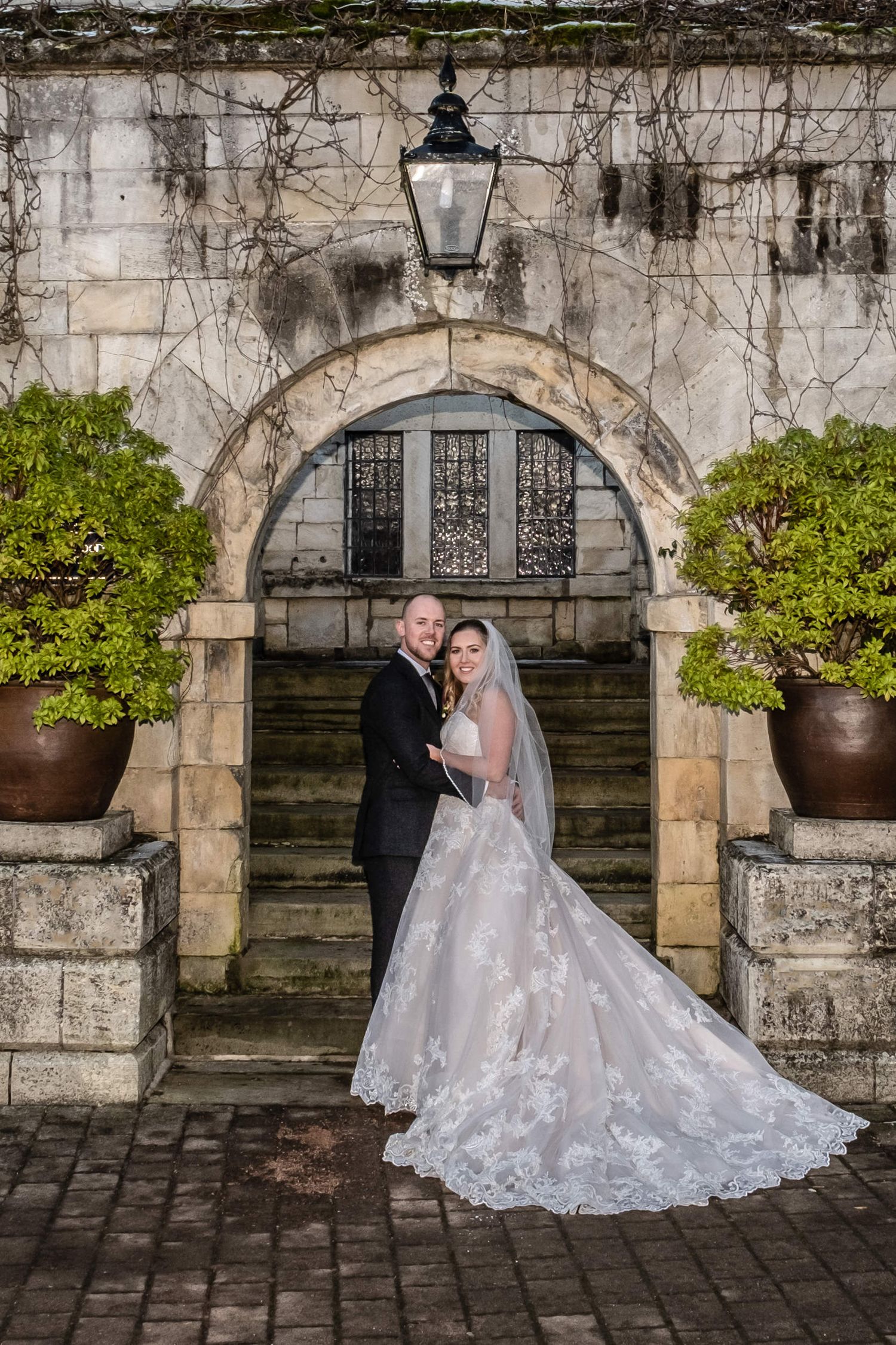 Elegant wedding portrait in front of stone archway with trailing vines and lantern.