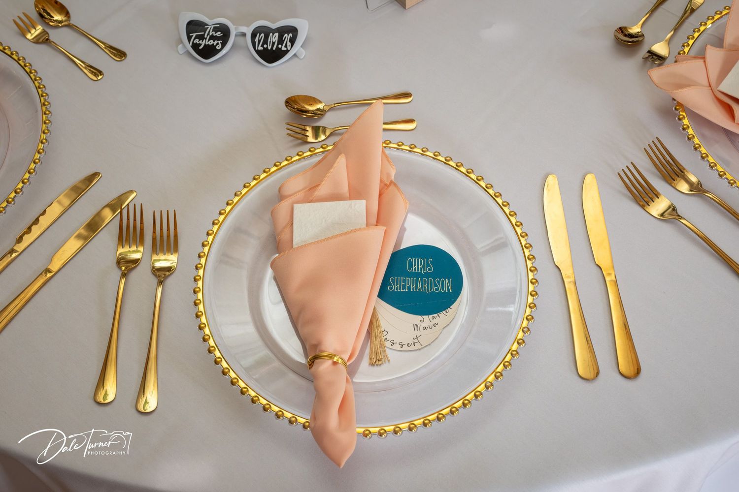Elegant wedding table setting with gold cutlery, beaded charger plate, peach napkin, and personalized place card.