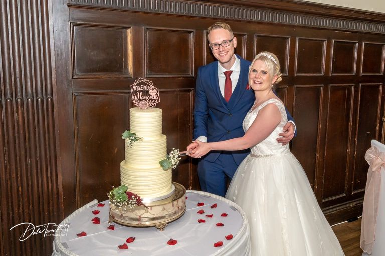Wedding reception setup showing elegant white cake and formal table settings in a grand wooden-paneled room.