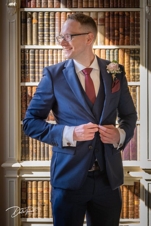 A formal wedding attire detail showing a navy suit with red tie against a backdrop of vintage library bookshelves.