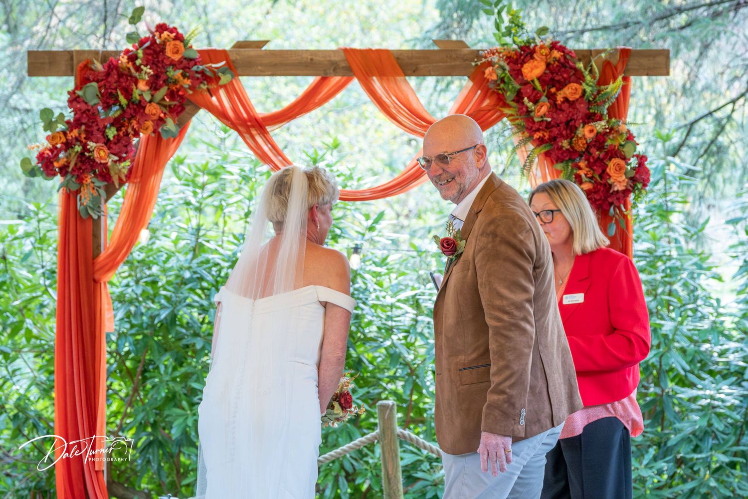 Wedding ceremony under a red floral arch with draped fabric in an outdoor garden setting.