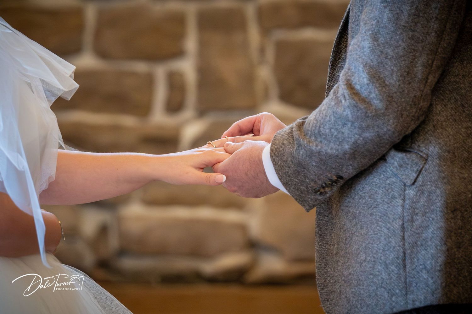 Close-up view of hands exchanging wedding rings during a romantic ceremony against a rustic brick wall backdrop.