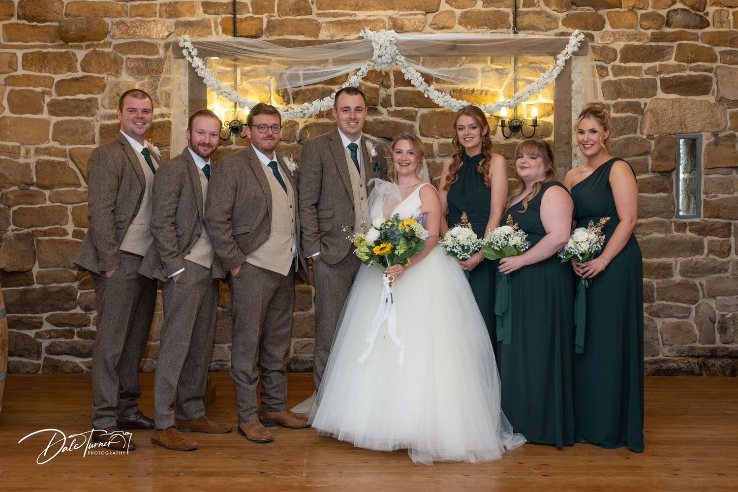 Wedding party poses together against rustic stone wall backdrop in formal attire.