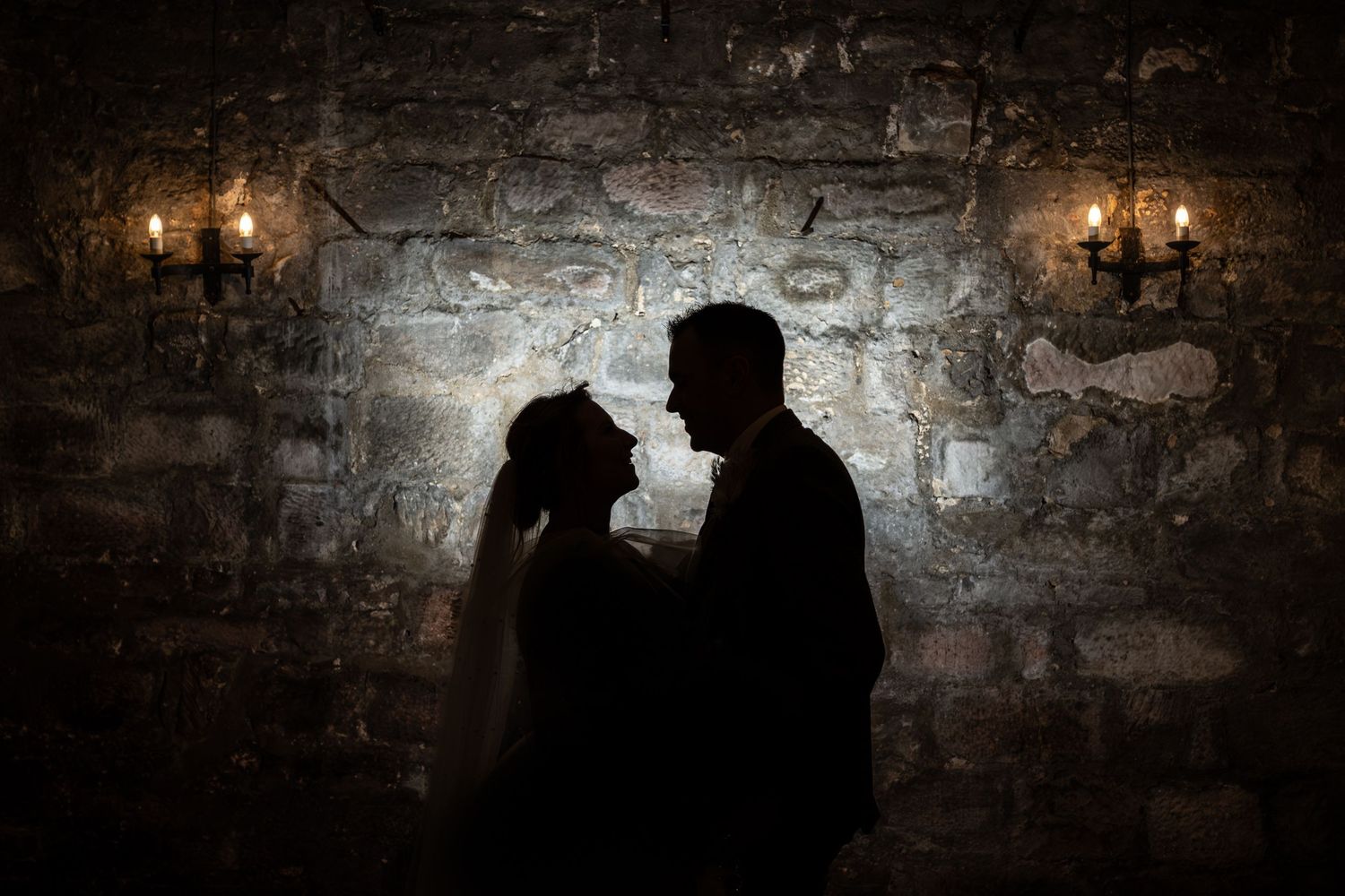Silhouetted couple stands intimately against rustic stone wall with vintage wall sconces glowing in the darkness.