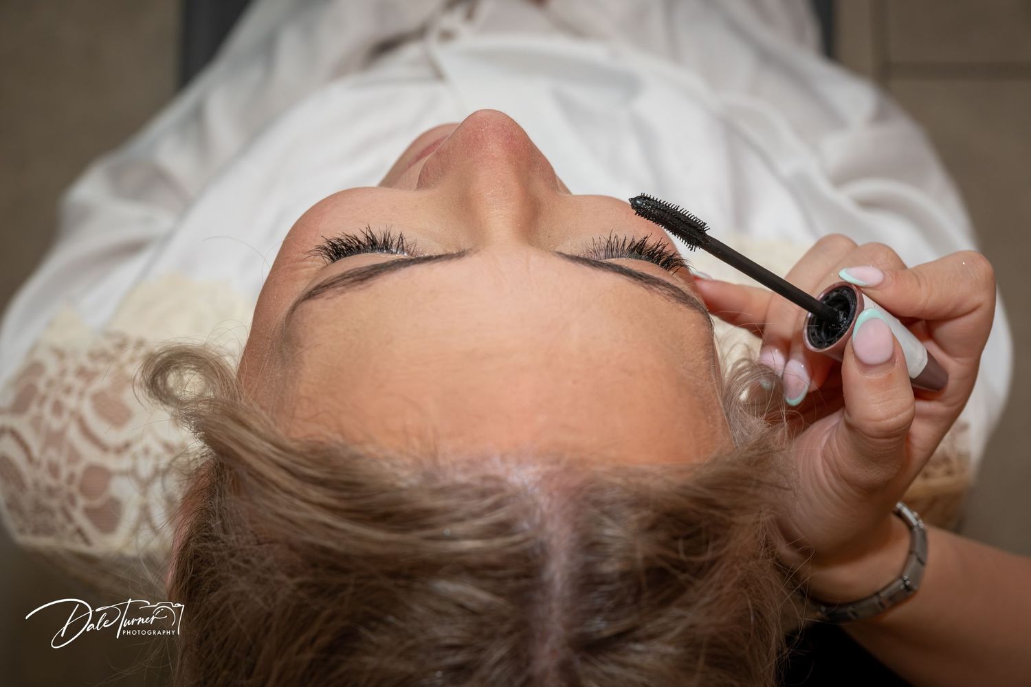 A makeup artist applies mascara to a bride in a white lace gown, captured from above in a professional bridal beauty session.
