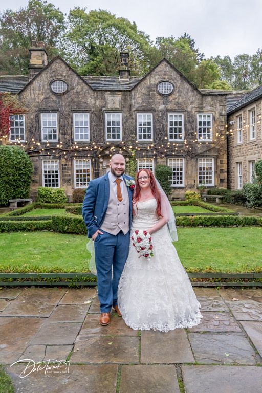 Wedding couple poses in front of a historic stone building with manicured gardens.