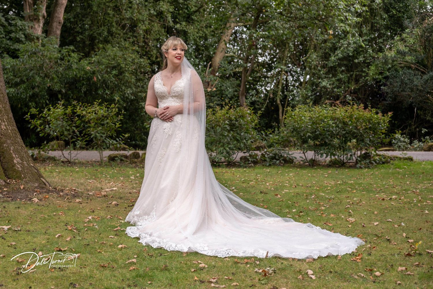 Person in lace wedding gown with long train poses in a garden setting.