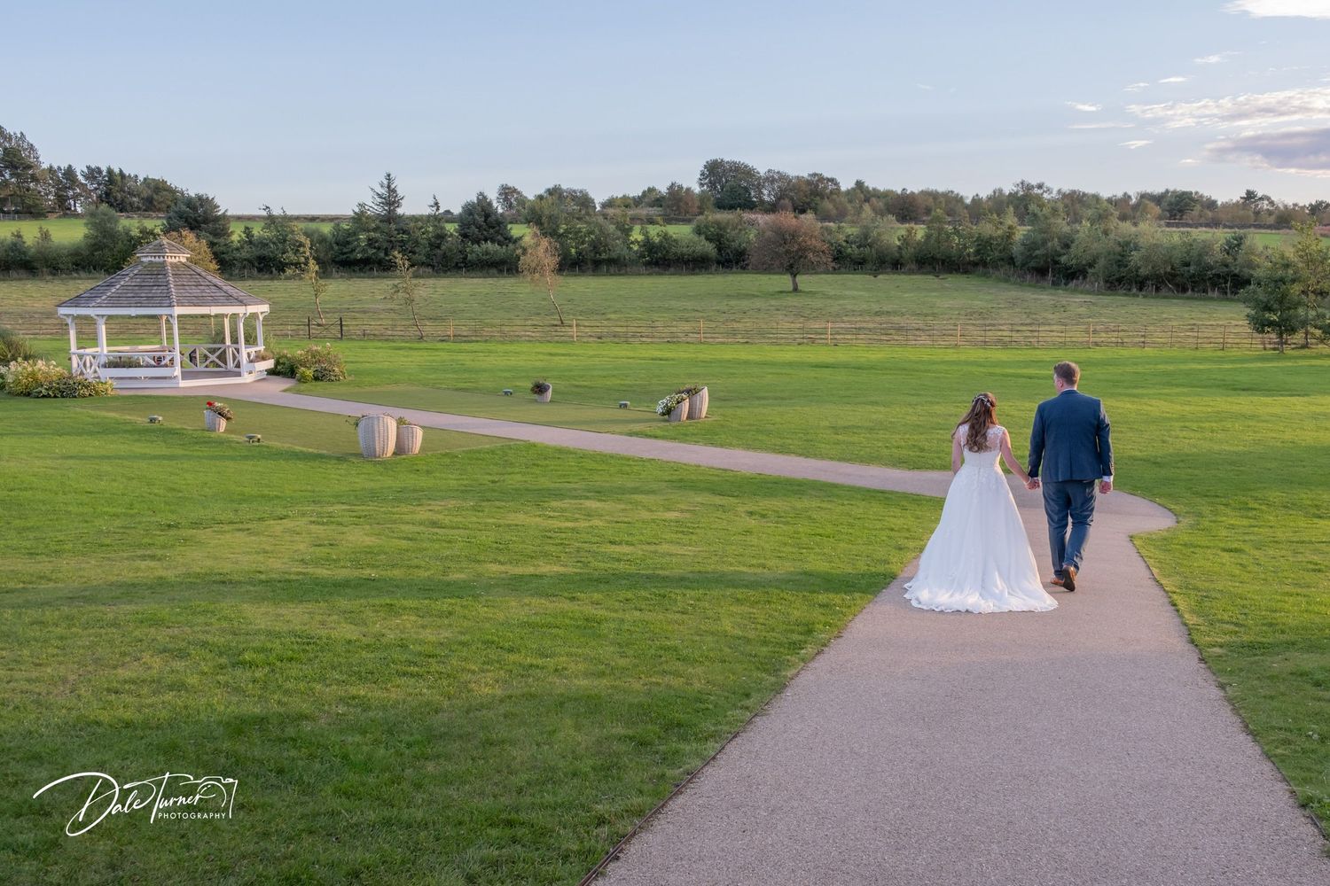 Wedding couple walking along a scenic pathway surrounded by open green fields.