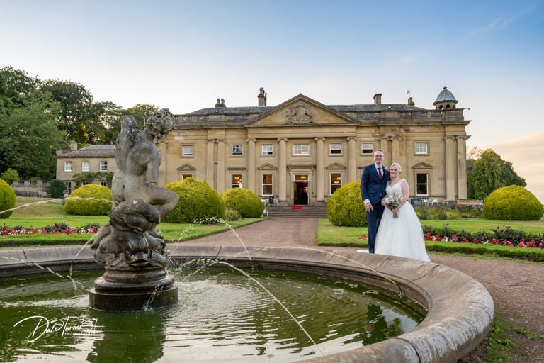 Wedding couple stands by ornate fountain in front of grand historic mansion.