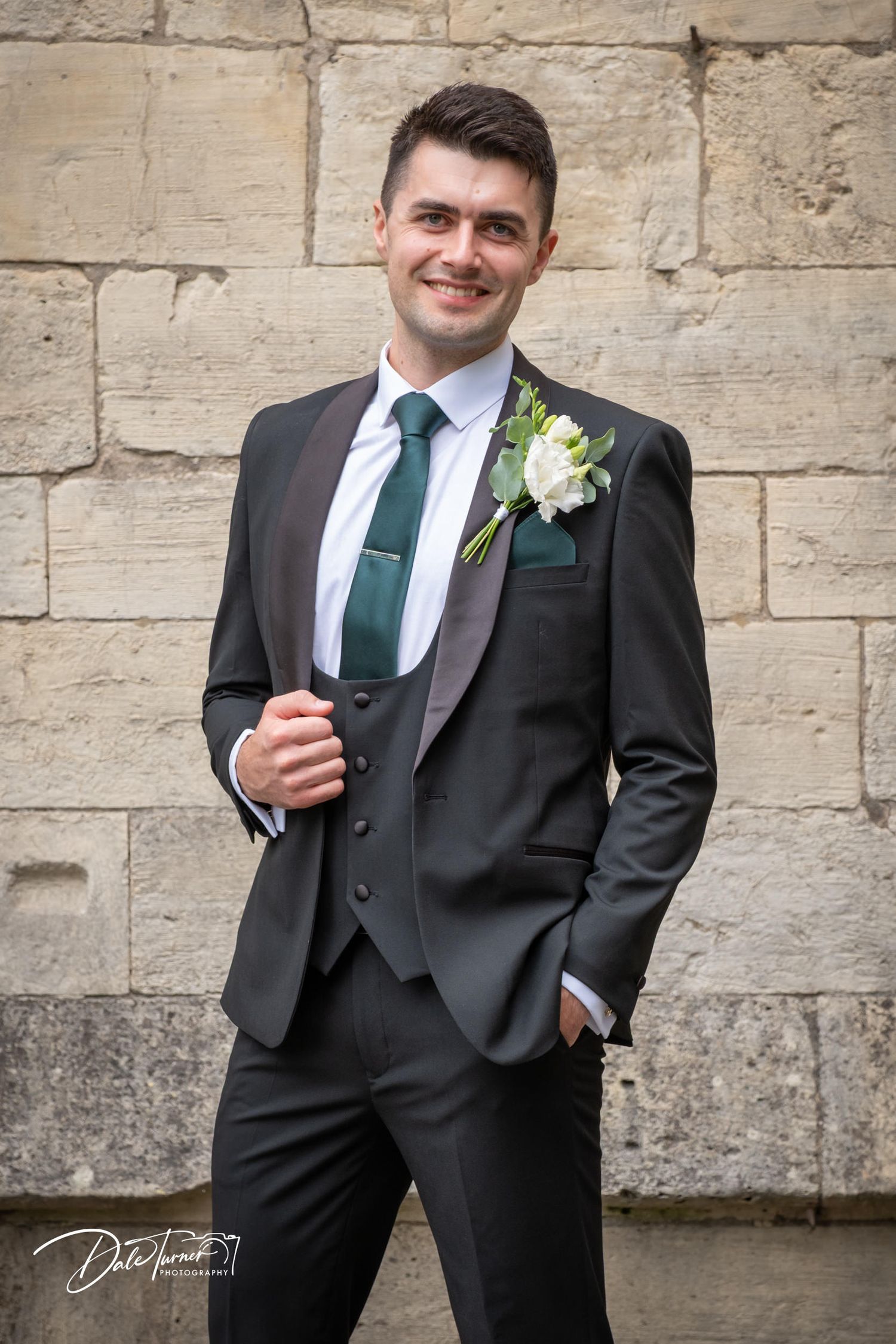 Groom in dark suit with green tie and white boutonniere poses against stone wall.