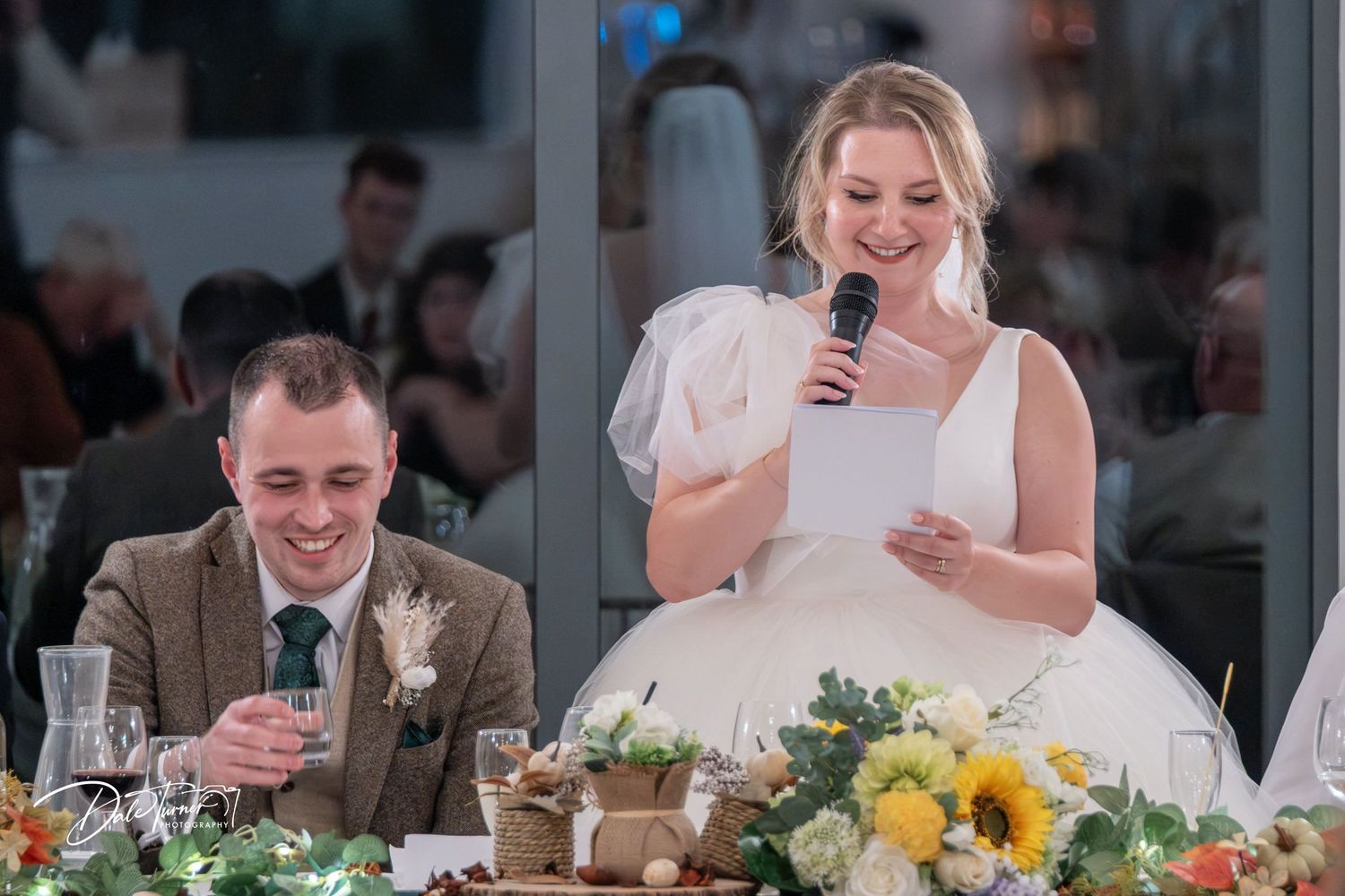 Wedding reception guests laugh and smile during emotional speeches at tables decorated with yellow sunflowers.
