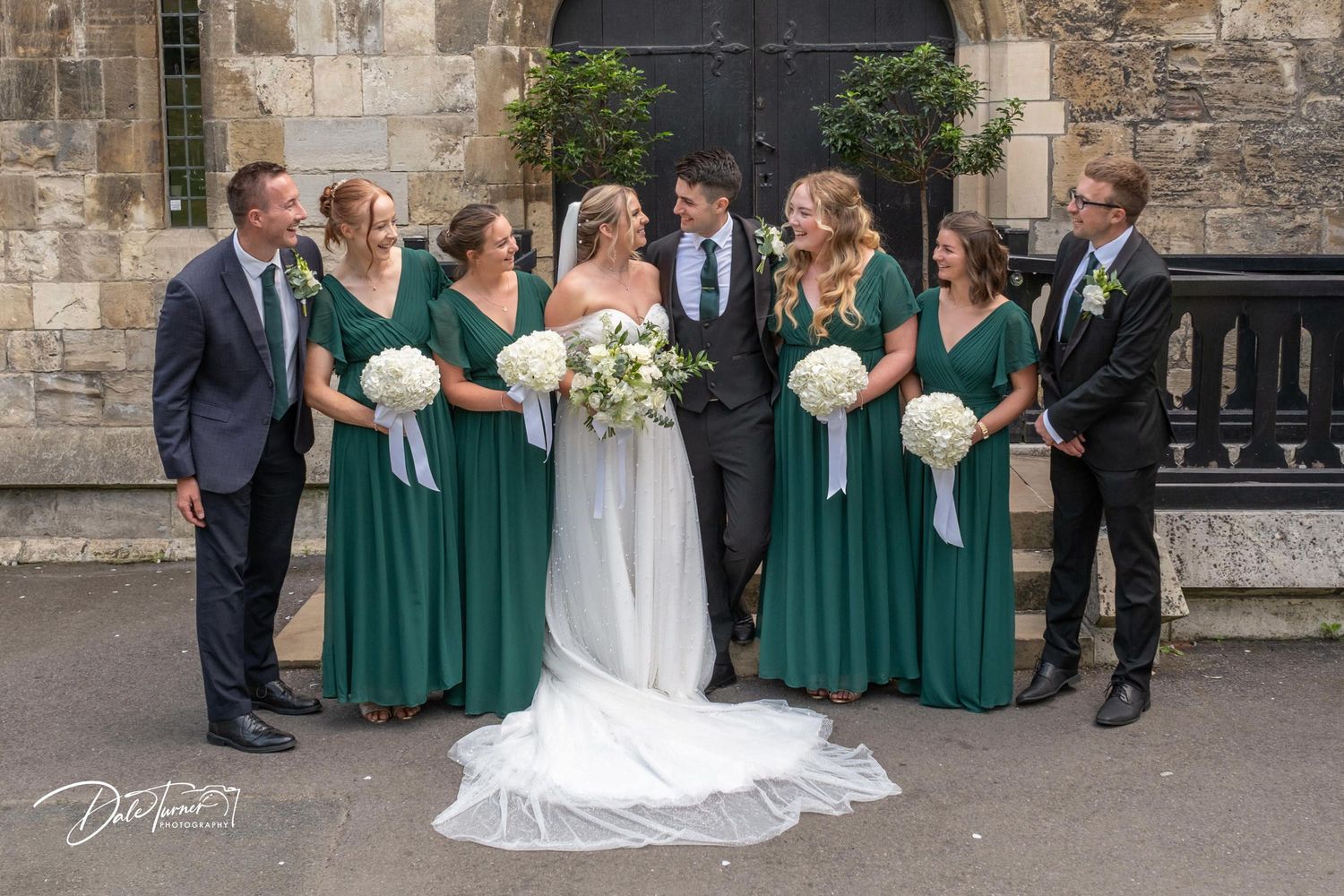 Wedding party lined up against stone wall for formal group photos with emerald green and white color scheme.