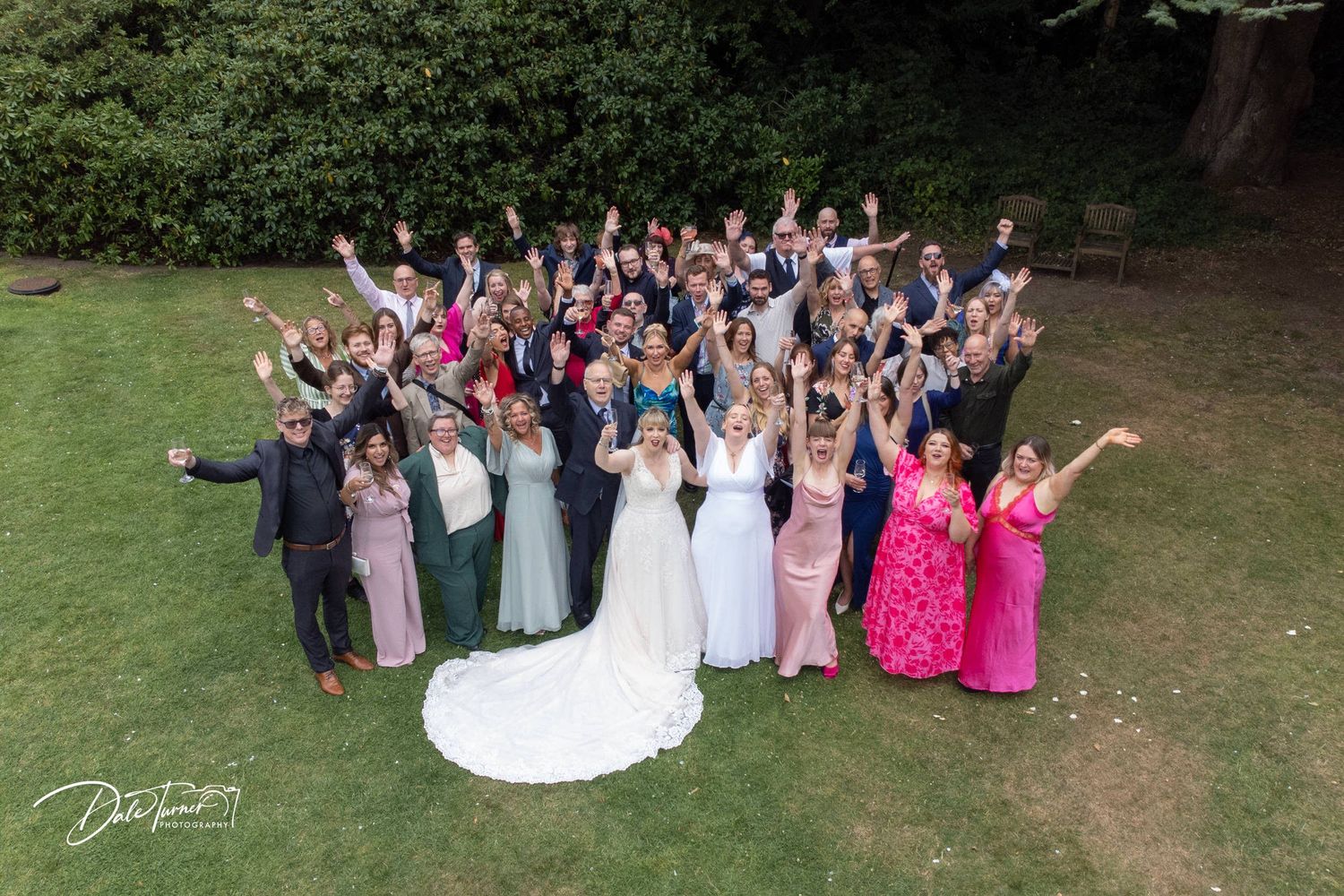 Large wedding group poses outdoors for formal photo with trees in background.