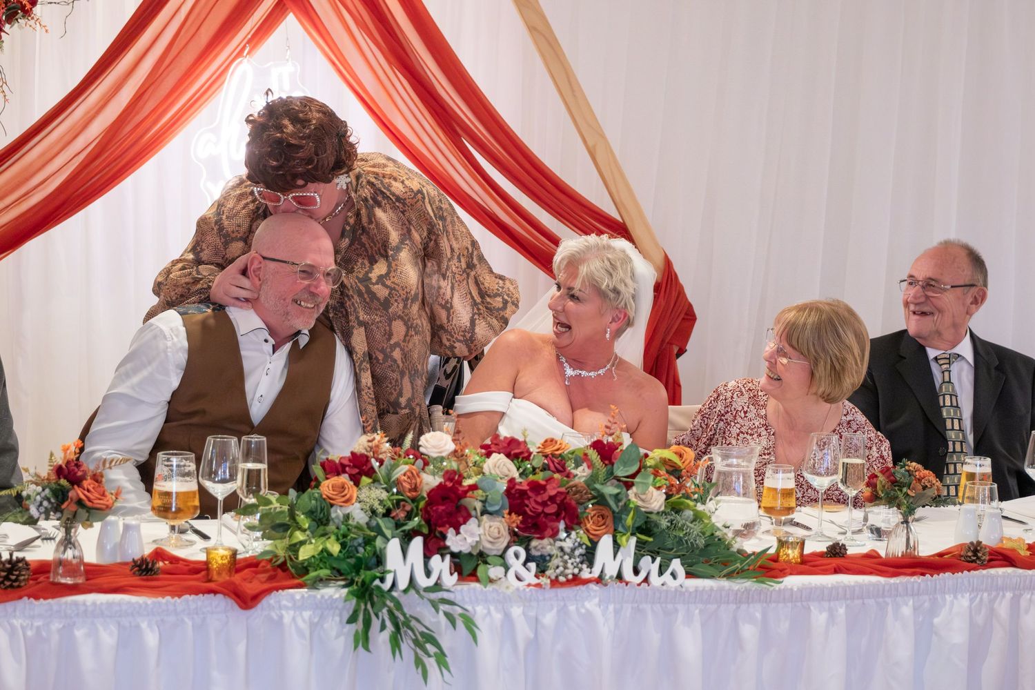Wedding reception head table with red and white decor under elegant draped fabric.