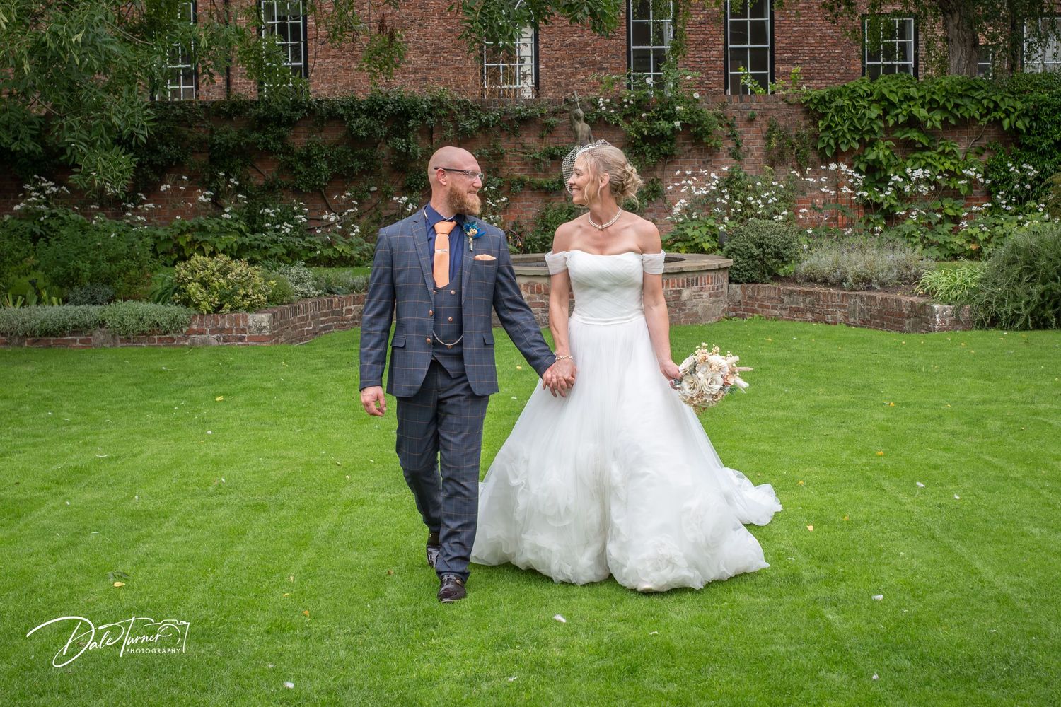 Bride and groom walking hand in hand on lush green lawn outside a brick manor house on their wedding day.
