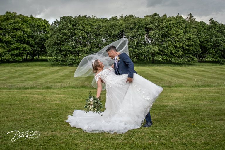 Newlyweds embrace on a spacious lawn surrounded by trees and natural scenery.
