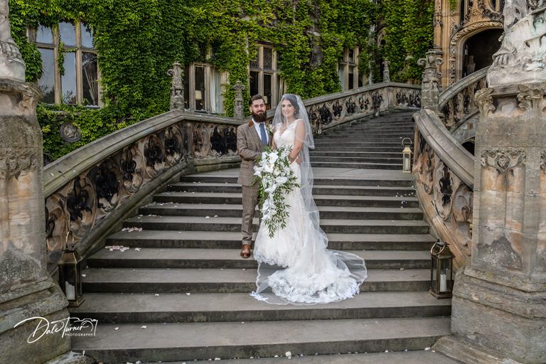 A couple stands on ornate stone steps covered in ivy at a grand estate.