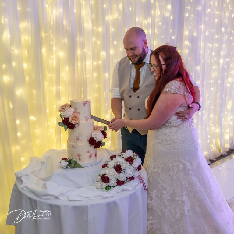 Couple cutting their elegant white and pink wedding cake in front of illuminated backdrop.