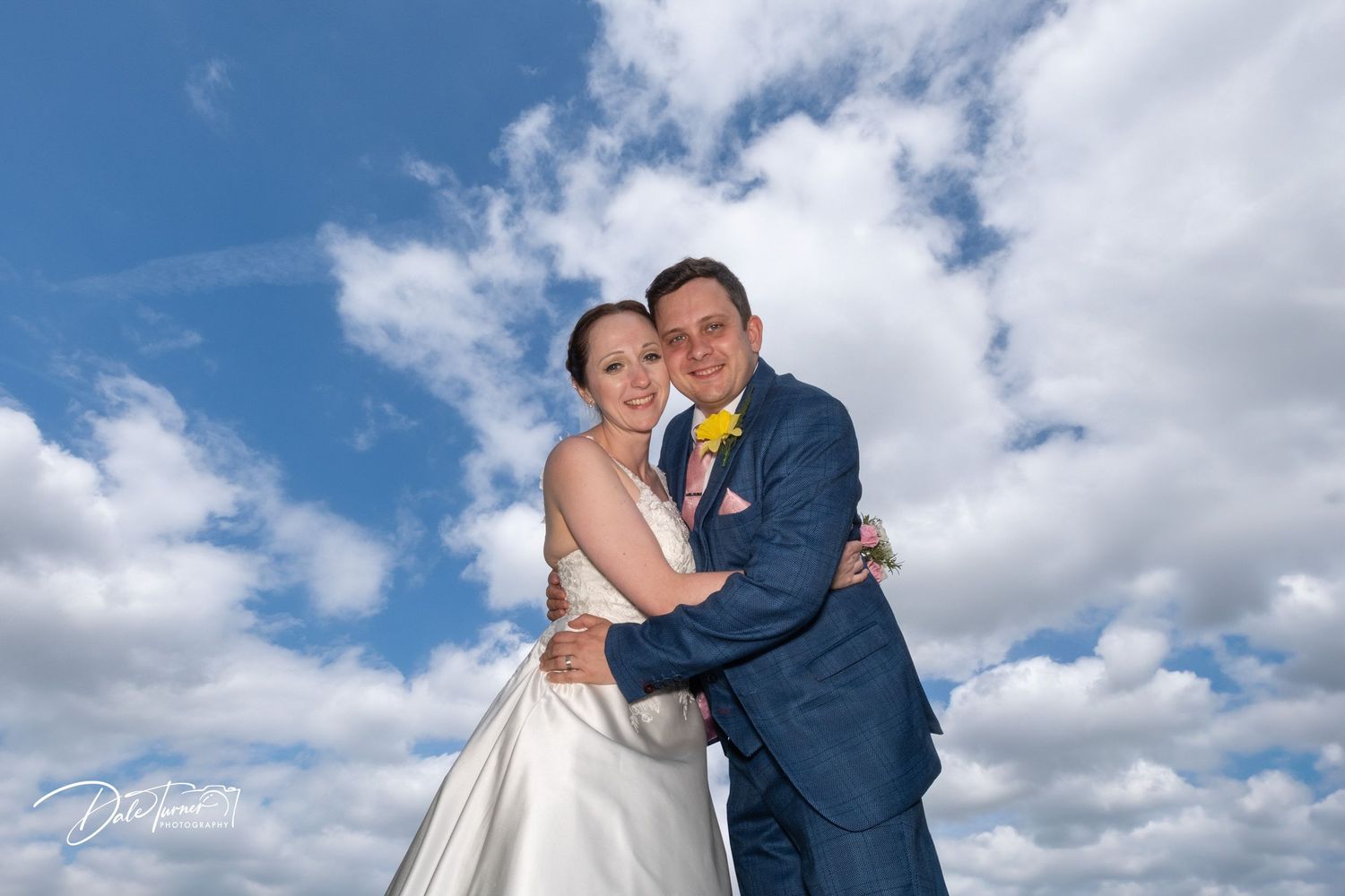 Wedding couple embraces under dramatic blue sky with white puffy clouds.