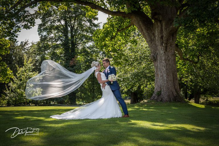 Wedding couple poses in scenic garden setting with flowing veil caught in gentle breeze.