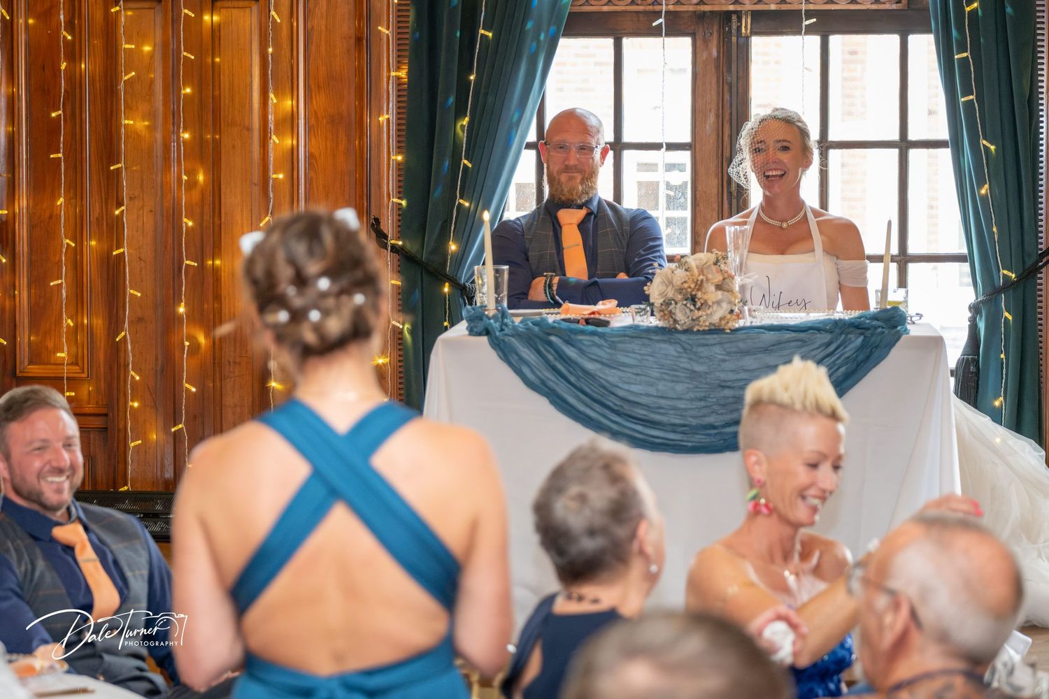 Bride and groom seated at sweetheart table during wedding reception with guests in blue attire in foreground.