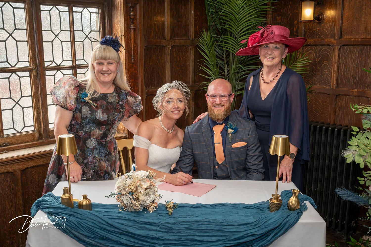 Bride and groom sign wedding register at ornate wooden table with two female witnesses in colorful hats and dresses.