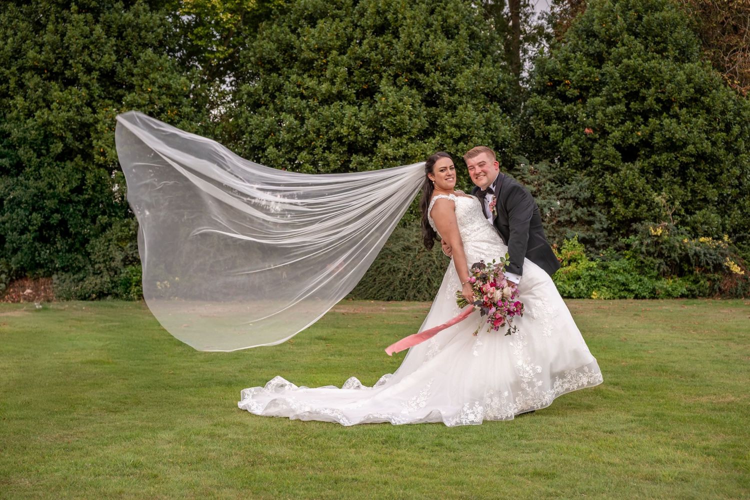 A flowing veil catches the breeze as a couple poses on manicured lawn grounds.